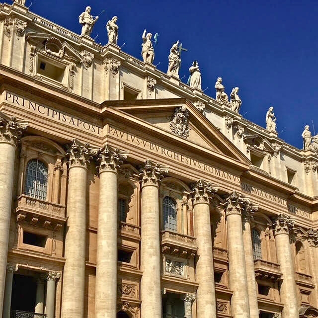 Fachada principal da Basílica de São Pedro, no Vaticano, com colunas e estátuas de Cristo e dos Apóstolos no alto da basílica. 