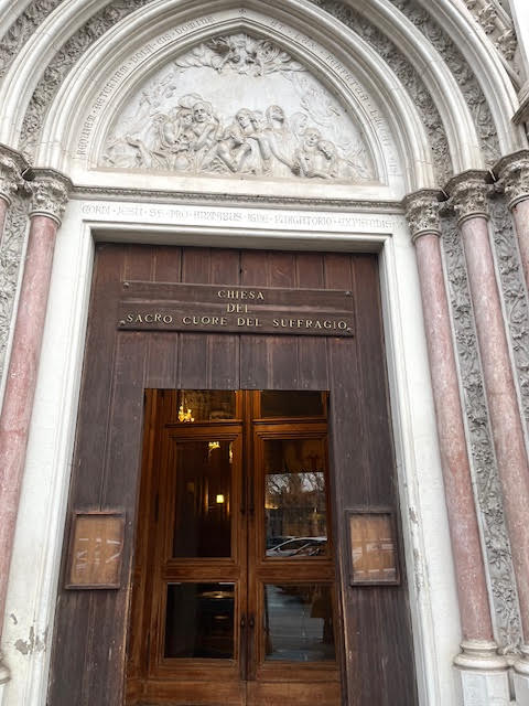 Fachada da Igreja do Sagrado Coração do Sufrágio em Roma, com o portal de madeira e esculturas em relevo sobre o frontão.