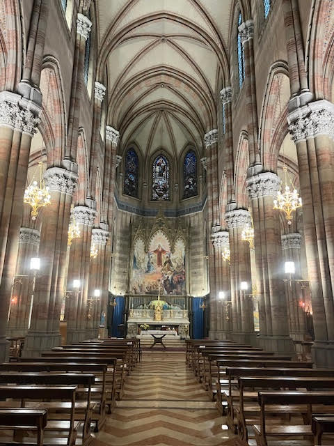 Interior da Igreja do Sagrado Coração do Sufrágio em Roma, com o altar principal, vitrais coloridos e colunas em estilo neogótico.