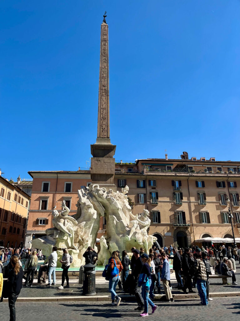 Fonte dos Quatro Rios na Piazza Navona, em Roma, com o obelisco central e visitantes ao redor em um dia ensolarado.