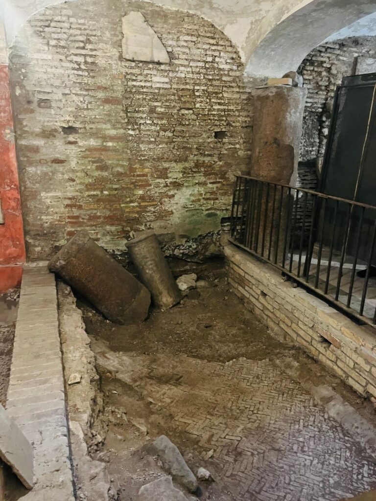 Ruínas arqueológicas sob a Basílica de Santa Cecília, com colunas romanas caídas e paredes de tijolos antigos que, segundo a tradição, pertencem à casa da santa.
