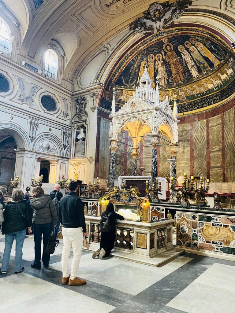 Interior da Basílica de Santa Cecília em Trastevere, com fiéis em oração diante do altar principal e da famosa escultura de Stefano Maderno representando a posição do corpo da santa.