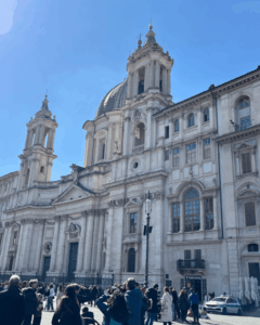 Fachada barroca da Igreja de Santa Inês em Agonia, em Roma, com céu azul fiéis em frente ao templo.