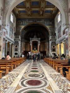 Vista do interior da Basílica de Santa Prassede, com o corredor central em mármore e o altar ao fundo, adornado por colunas e mosaicos bizantinos.