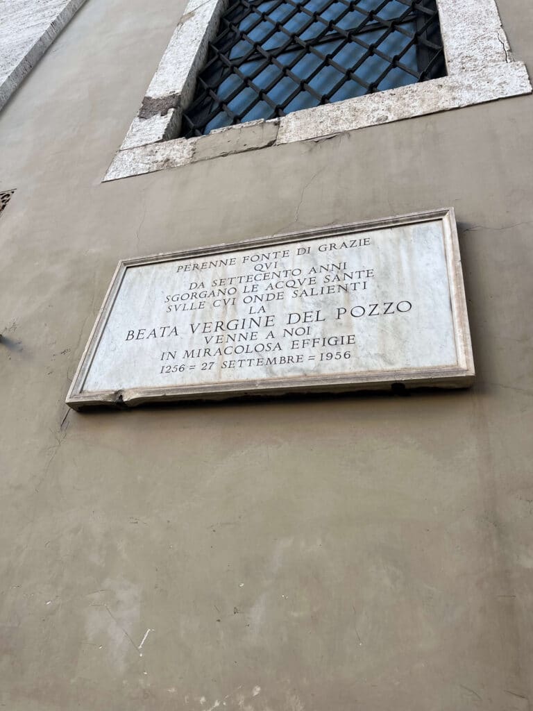 Placa comemorativa dos 700 anos do milagre da Madonna del Pozzo, na fachada externa de Santa Maria in Via, com a inscrição em italiano datada de 1256-1956