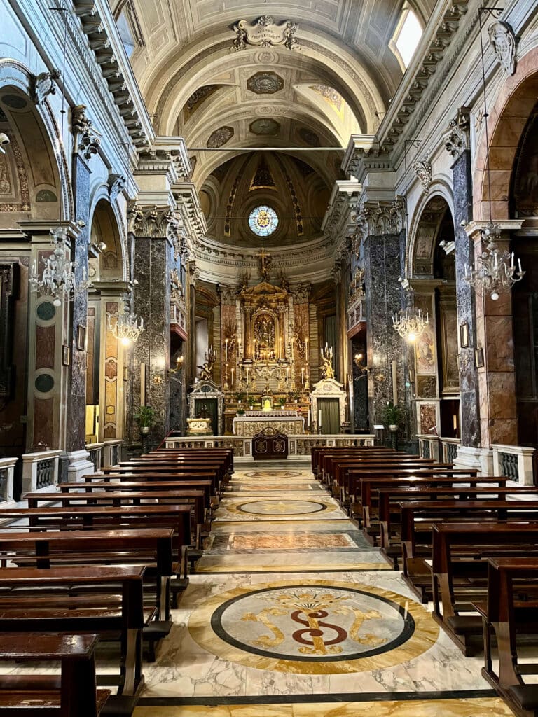 Nave central da Igreja de Santa Maria in Via, em Roma, vista em direção ao altar-mor dourado, com bancos de madeira e piso decorado em mármore.