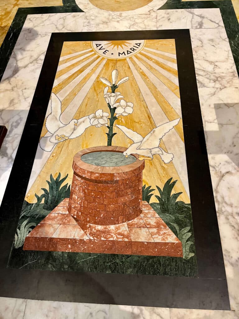 Painel em opus sectile no chão da Capela da Madonna del Pozzo em Santa Maria in Via, representando o poço, duas pombas e um lírio sob a inscrição "Ave Maria"